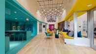 Located at 10920 Garfield Ave in South Gate, Los Angeles, this single-floor space makes a striking first impression with its bold, contemporary design language. The interior is defined by a vivid palette of teal, yellow, and magenta, applied to architectural elements, accent walls, and furnishings throughout, creating an energetic and welcoming atmosphere.

The main lobby and lounge area features soaring ceilings adorned with a dramatic, large-scale pendant light installation — dozens of globe lights suspended from a geometric black grid — that serves as a central design focal point. Light hardwood flooring runs throughout, lending warmth and continuity to the space. A built-in teal reception counter anchors one side, framed by deep blue abstract artwork. On the opposite side, a generous built-in bench with a yellow upholstered seat and an eye-catching herringbone wood accent wall provides ample seating.

Colorful lounge chairs in teal, yellow, and magenta are arranged throughout for casual gathering. Floor-to-ceiling glass walls and doors allow abundant natural light to pour in, connecting the interior to outdoor views. A dedicated mailroom area features a clean white counter, bold magenta accents, vibrant pendant lights, and a wall of resident mailboxes accented with lush greenery. Additional glass-enclosed rooms provide flexible spaces for meetings or focused activities.