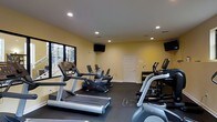 This fitness center offers a practical and functional workout environment designed to support a variety of exercise routines. The room features warm yellow walls and a dark rubberized flooring, providing a clean contrast that is both visually appealing and purpose-built for athletic use. Recessed lighting throughout the ceiling ensures the space is well-lit, creating a bright and energizing atmosphere conducive to exercise.

The cardio section is well-stocked, featuring treadmills, elliptical trainers, and a stationary bike, all positioned to allow comfortable use without overcrowding. A dumbbell rack is visible along one wall, offering options for strength training. Multiple flat-screen televisions are mounted on the walls, keeping users entertained during workouts.

A large window along one wall allows natural light to filter into the space, adding openness to the room. A standard door provides access to and from the facility. The overall layout is thoughtfully organized, balancing cardio and strength equipment efficiently within the available space, making it a well-rounded fitness facility suitable for everyday use.