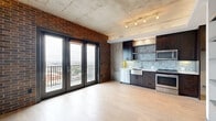 This single-floor urban loft masterfully blends industrial character with modern sophistication. The open-plan living area is defined by striking exposed brick walls, raw concrete ceilings with track lighting, and light hardwood floors that run throughout. Expansive black-framed French doors flood the space with natural light and open directly onto a balcony offering panoramic city views, creating a seamless indoor-outdoor connection.

The kitchen is well-appointed with dark espresso cabinetry, crisp white quartz countertops, a deep farmhouse sink, and stainless steel appliances. A mosaic glass tile backsplash adds texture and visual interest, complementing the adjacent exposed brick.

The bedroom features the same soaring concrete ceiling and includes custom built-in shelving with a media niche and dark wood cabinetry, providing generous storage while maintaining the space's refined industrial aesthetic. A concrete accent wall adds architectural depth.

The bathroom is finished with large-format tile flooring, a soaking tub with vertically stacked white subway tile surround, dark vanity cabinetry with quartz countertops, and contemporary sconce lighting. A hallway connects all areas with clean lines, smooth walls, and a bold paneled entry door, tying together the industrial-chic design language found throughout the entire space.
