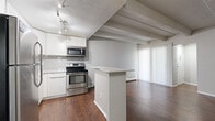 This 2-bedroom, 1-bathroom unit is thoughtfully laid out across a single floor, offering a functional and cohesive living environment. The kitchen is a standout feature, equipped with stainless steel appliances including a refrigerator, electric range, and over-the-range microwave. White cabinetry with a clean, modern profile pairs with white subway tile backsplash and light countertops to create a bright, contemporary cooking space. A peninsula counter provides a natural division between the kitchen and the adjoining living area, which features rich, dark wood-style flooring and exposed beam ceilings that add architectural character. Vertical blinds cover a large window or sliding door in the living room, allowing for adjustable natural light. Baseboard heating units are present throughout the unit. The bedrooms are carpeted for comfort, with one bedroom showcasing a wide double sliding-door closet that maximizes storage. The bathroom features a dark-toned vanity with a white countertop and light tile flooring, connected to the unit via a hallway. The overall color palette is neutral and clean, with textured walls and ceilings contributing to the unit's distinct character.