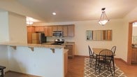 This two-story residence offers a thoughtfully designed layout that blends functionality with comfortable living. The open-concept kitchen is a standout feature, showcasing warm-toned wood cabinetry, granite countertops, and a full suite of stainless steel appliances including a gas range, microwave, and refrigerator. A peninsula with breakfast bar seating extends into the living area, creating a natural gathering space ideal for casual dining or entertaining. Recessed lighting illuminates the kitchen, complemented by a decorative pendant light fixture that anchors the adjacent dining area. The dining space is cozy yet open, furnished with a wood-top table and classic black chairs set atop a patterned area rug, with a multi-panel mirror on the wall adding depth and light to the room. Warm hardwood flooring runs throughout the main level, tying the spaces together cohesively. The home spans two floors, offering separation between living and sleeping areas. The overall ambiance is clean, bright, and welcoming, with neutral wall tones that provide a versatile backdrop for a variety of interior styles. This space is well-suited for those seeking a move-in ready home with practical, modern finishes.