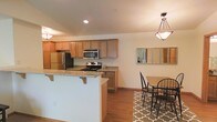 This two-story residence offers a thoughtfully designed layout that blends functionality with comfortable living. The open-concept kitchen is a standout feature, showcasing warm-toned wood cabinetry, granite countertops, and a full suite of stainless steel appliances including a gas range, microwave, and refrigerator. A peninsula with breakfast bar seating extends into the living area, creating a natural gathering space ideal for casual dining or entertaining. Recessed lighting illuminates the kitchen, complemented by a decorative pendant light fixture that anchors the adjacent dining area. The dining space is cozy yet open, furnished with a wood-top table and classic black chairs set atop a patterned area rug, with a multi-panel mirror on the wall adding depth and light to the room. Warm hardwood flooring runs throughout the main level, tying the spaces together cohesively. The home spans two floors, offering separation between living and sleeping areas. The overall ambiance is clean, bright, and welcoming, with neutral wall tones that provide a versatile backdrop for a variety of interior styles. This space is well-suited for those seeking a move-in ready home with practical, modern finishes.
