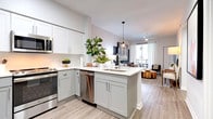 This single-floor residence showcases a thoughtfully designed open-concept layout finished with light wood-look flooring and crisp white walls throughout. The kitchen is outfitted with grey shaker-style cabinetry, white quartz countertops, a peninsula with an undermount sink, and a full suite of stainless steel appliances including a French door refrigerator, gas range, built-in microwave, and dishwasher. A pendant light over the peninsula provides both task lighting and visual interest.

The living area flows seamlessly from the kitchen and dining space, offering room for comfortable seating and a ceiling fan overhead. Large windows and a sliding glass door allow natural light to fill the space and provide access to a private balcony. An accent wall adds a bold design element to the living zone.

The bedroom features a ceiling fan, natural light from a window, a walk-in closet, and a teal accent wall that creates a distinctive focal point. The bathroom includes a tub-shower combo with subway tile, a grey vanity with white countertop, and an illuminated mirror.

A dedicated home office nook includes a built-in desk area with convenient access to an in-unit laundry closet housing a full-size washer and dryer.