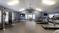 This single-level fitness center offers a generous, open workout environment designed to accommodate a comprehensive range of exercise equipment. The room's most striking architectural feature is its vaulted ceiling, which rises to a notable peak along the center and is accented by a pendant ceiling fan, lending the space a feeling of openness well beyond its footprint. Blue-gray painted walls and crisp white trim create a clean, focused atmosphere throughout.

The flooring consists of durable rubber matting, providing both cushioning and traction suitable for high-impact exercise. A prominent bay window along one wall allows natural light to filter into the space, complemented by flush fluorescent light fixtures mounted along the ceiling slopes. Multiple wall-mounted flat-screen televisions are positioned for visibility across different workout zones.

The space is well-organized with distinct areas for cardiovascular and strength training. Cardio equipment includes several treadmills, elliptical trainers, and a stationary bike. A large framed wall mirror along one section enhances the sense of depth. Strength training is supported by a cable machine, a weight bench, a dumbbell rack, a power tower, and a heavy bag. Two white panel doors provide entry and exit access, completing this fully functional, thoughtfully appointed fitness facility.