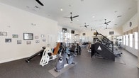 This fitness center is a spacious, single-floor facility designed to support a full range of workout routines. The interior features durable rubber flooring throughout, providing both safety and comfort during exercise. Crown molding and crisp white walls lend a clean, polished aesthetic that sets this gym apart from typical fitness spaces.

The facility is divided into distinct workout zones. A strength training area houses a robust selection of Cybex resistance machines, including chest press, leg press, and cable systems, alongside a dedicated free weight section with a full rack of dumbbells and adjustable benches. A separate cardio zone features stationary bikes, treadmills, stair climbers, and other aerobic equipment.

Natural light fills the space through large floor-to-ceiling windows, which offer views of an outdoor pool and courtyard area. Wall-mounted mirrors amplify the sense of openness, while multiple ceiling fans and recessed lighting ensure a comfortable environment. Mounted televisions are positioned throughout for entertainment during workouts.

Motivational artwork and framed photography decorate the walls, contributing to an energizing atmosphere. The thoughtful layout provides ample room to move between stations, making this fitness center a functional and inviting space for daily exercise.