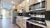 This well-appointed single-floor unit offers a thoughtfully designed layout that balances function and modern style throughout. The kitchen is a standout feature, showcasing light maple cabinetry with sleek black hardware, a dark subway tile backsplash, white quartz countertops, and a full suite of stainless steel appliances including a refrigerator, electric range, over-the-range microwave, and dishwasher. Recessed lighting illuminates the space, while wood-style plank flooring runs continuously throughout the unit, lending warmth and visual cohesion.

The open living and dining area provides generous, flexible space with soft sage-toned walls and clean white baseboards. A sliding glass door leads directly to a private balcony with a metal railing, offering outdoor views of mature trees and bringing natural light into the room. An exposed metal ventilation duct adds an industrial design accent overhead.

The bathroom is finished with the same light maple cabinetry seen in the kitchen, featuring a single vanity with an integrated sink, a large mirror, multi-bulb lighting fixture, and grab bars. The overall floor plan, visible in the 3D model, reveals a compact yet efficient layout with distinct zones for living, cooking, and private use — all connected by a clean, modern design aesthetic.