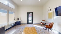 This single-floor business center is thoughtfully designed to support productivity and collaboration in a contemporary, welcoming environment. The space is characterized by soaring ceilings, warm neutral walls, and light wood-tone flooring that runs throughout, creating a bright and cohesive aesthetic.

Upon entry through a sleek black-framed glass door, visitors are greeted by an open reception area furnished with a tufted cognac leather bench atop a stylish patterned area rug. Adjacent to the entry, a dedicated work zone features a built-in white countertop workstation flanked by dark cabinetry, set beneath a wide window that draws in abundant natural light — ideal for focused individual work.

A small meeting area accommodates intimate discussions, furnished with a glass-top table, two tan leather chairs, a wall-mounted display, and accent lighting. Artwork adorns the walls, adding a curated touch to the professional atmosphere. Deeper into the space, a well-appointed kitchenette is visible, outfitted with cabinetry and pendant lighting, providing convenience for extended visits.

Large windows along the length of the space ensure consistent daylighting throughout. The clean lines, bold black accents, and modern furnishings come together to create a polished, functional environment well-suited for work and collaboration.