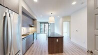 This well-designed two-bedroom, one-bathroom unit offers a thoughtful open-concept layout with light wood-tone flooring throughout and a clean, contemporary aesthetic. The kitchen is a standout feature, outfitted with full-height textured cabinetry in warm gray tones, a striking dark granite waterfall island with built-in shelving, white subway tile backsplash, and a complete suite of stainless steel appliances including a refrigerator, dishwasher, and range. Recessed lighting and a pendant fixture above the island enhance the space's modern appeal. The living area flows naturally from the kitchen and opens to a private balcony through black-framed sliding glass doors, offering views of the surrounding urban streetscape. Both bedrooms feature light wood flooring, crisp white walls, and generous natural light — the primary bedroom includes a large window with city views. The bathroom is finished with a wide double-sink vanity, dark stone countertop, textured gray cabinetry, and a backlit mirror, lending a spa-like quality. A dedicated closet provides additional storage. The unit's cohesive, modern finishes and efficient layout make it a practical and stylish living space.