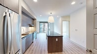 This well-designed two-bedroom, one-bathroom unit offers a thoughtful open-concept layout with light wood-tone flooring throughout and a clean, contemporary aesthetic. The kitchen is a standout feature, outfitted with full-height textured cabinetry in warm gray tones, a striking dark granite waterfall island with built-in shelving, white subway tile backsplash, and a complete suite of stainless steel appliances including a refrigerator, dishwasher, and range. Recessed lighting and a pendant fixture above the island enhance the space's modern appeal. The living area flows naturally from the kitchen and opens to a private balcony through black-framed sliding glass doors, offering views of the surrounding urban streetscape. Both bedrooms feature light wood flooring, crisp white walls, and generous natural light — the primary bedroom includes a large window with city views. The bathroom is finished with a wide double-sink vanity, dark stone countertop, textured gray cabinetry, and a backlit mirror, lending a spa-like quality. A dedicated closet provides additional storage. The unit's cohesive, modern finishes and efficient layout make it a practical and stylish living space.