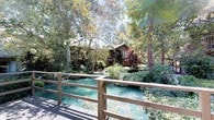 This single-story property offers a truly distinctive living experience defined by its deep integration with the natural landscape surrounding it. Mature trees canopy the grounds, filtering sunlight and creating a sense of seclusion rarely found in residential settings. A tranquil pond with clear, turquoise-tinted water winds through the property, spanned by a wooden footbridge with a simple railing that connects outdoor areas and invites leisurely strolling through the lush, carefully cultivated grounds. Ornamental plantings, including a striking Japanese maple, add layers of color and texture throughout the landscape.

The home's exterior reflects a rustic, organic aesthetic, with wood-clad siding and a deck or balcony that extends the living space outward toward the natural surroundings. Entry to the home is marked by a covered porch with aggregate concrete flooring and vertical wood paneling. A set of bold, warm-toned orange French doors serves as the focal point of the entryway, complemented by a classic geometric lantern-style wall sconce that adds a touch of elegance. The textured ceiling and understated finishes of the entry set a relaxed, welcoming tone that carries throughout the home.