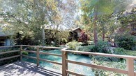This single-story property offers a truly distinctive living experience defined by its deep integration with the natural landscape surrounding it. Mature trees canopy the grounds, filtering sunlight and creating a sense of seclusion rarely found in residential settings. A tranquil pond with clear, turquoise-tinted water winds through the property, spanned by a wooden footbridge with a simple railing that connects outdoor areas and invites leisurely strolling through the lush, carefully cultivated grounds. Ornamental plantings, including a striking Japanese maple, add layers of color and texture throughout the landscape.

The home's exterior reflects a rustic, organic aesthetic, with wood-clad siding and a deck or balcony that extends the living space outward toward the natural surroundings. Entry to the home is marked by a covered porch with aggregate concrete flooring and vertical wood paneling. A set of bold, warm-toned orange French doors serves as the focal point of the entryway, complemented by a classic geometric lantern-style wall sconce that adds a touch of elegance. The textured ceiling and understated finishes of the entry set a relaxed, welcoming tone that carries throughout the home.