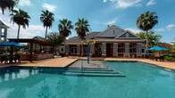 Located at 15270 Voss Rd in Sugar Land, Texas, this single-story community clubhouse and amenity center at Avana Sugar Land offers a well-appointed outdoor retreat designed for recreation and relaxation. The building's exterior showcases a classic combination of warm red brick and gray lap siding, complemented by large floor-to-ceiling windows and white-trimmed columns that lend a polished, welcoming aesthetic.

The centerpiece of the space is a sparkling resort-style swimming pool, complete with splash pad features and stainless steel railings, set atop a generously sized paver-stone deck. Swaying palm trees frame the pool area, enhancing the relaxed, vacation-inspired atmosphere. Sun loungers and bright blue umbrellas are arranged poolside, providing ample space for outdoor leisure under the Texas sky.

A covered porch and patio area runs along the building's exterior, offering shaded outdoor space for socializing. A concrete ping-pong table branded with the Avana Sugar Land name serves as a standout recreational feature on the patio. Expansive glass panels and large windows throughout ensure a seamless visual connection between the interior and the vibrant outdoor areas. The secured entry features a glass panel gate with electronic access, ensuring both safety and convenience for residents.