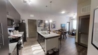 This single-floor residence showcases a thoughtfully designed open-concept layout that connects the kitchen, dining, and living areas into one cohesive and functional space. The kitchen serves as the centerpiece, anchored by a large granite-topped island with a double sink and pendant lighting that draws the eye upward toward the elevated ceilings. Dark cabinetry with stainless steel appliances, including a refrigerator and range, adds a sleek, contemporary aesthetic. The kitchen island also features bar seating, making it an ideal space for casual dining or entertaining. Wide-plank hardwood-style flooring runs throughout, unifying the entire living area with warmth and continuity. The dining area transitions naturally from the kitchen, offering a dedicated space for a full dining table and seating. Beyond it, the living area provides a comfortable retreat with ample natural light streaming in through well-dressed windows, complemented by a ceiling fan overhead. Neutral tones on the walls and thoughtfully placed wall art contribute to a clean, modern ambiance throughout. The arched interior doors add a subtle architectural detail that enhances the overall design.