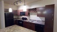 Located at 1200 E Parmer Lane in Austin, Texas, this single-floor unit offers a thoughtfully designed layout with a cohesive interior aesthetic throughout. The kitchen serves as a functional centerpiece, featuring rich dark-stained wood cabinetry, granite-style countertops, and black appliances including a refrigerator, electric range, microwave, and dishwasher. A built-in wine rack adds a distinctive touch, while pendant lighting and track lighting illuminate the space effectively. A kitchen island with matching countertop provides additional prep space and flows naturally into the living area. The living room is finished with warm hardwood-style flooring and includes a ceiling fan, with natural light filtering in through window blinds. The bedroom offers plush carpeting, a ceiling fan, and a window that brings in natural light, creating a comfortable retreat. A hallway connects the bedroom to the bathroom, which is appointed with a dark wood vanity, granite-style countertop, a large mirror, and a tub/shower combination. Neutral wall tones and consistent trim work throughout the unit contribute to a clean, uniform appearance that is both practical and inviting.