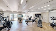 This fitness center is thoughtfully designed across two interconnected exercise rooms, offering a comprehensive workout environment with a clean, modern aesthetic. The space features striking herringbone-patterned wood-look flooring throughout, crisp white walls, and exposed beam ceilings fitted with industrial-style fluorescent lighting that keeps the environment bright and energizing.

Natural light flows generously through multiple large sliding glass doors and windows, providing views of an outdoor greenspace and creating an open, airy atmosphere. One room is anchored by a textured stone accent wall mounted with a large flat-screen television, adding a stylish focal point alongside an array of cardio equipment including ellipticals, a treadmill, a stair climber, and an air bike. A free weights rack with a flat bench rounds out the strength training options.

The second room expands the offering with a multi-station cable machine, additional benches, and further cardio equipment, making it well-suited for varied workout routines. Mirrored walls in sections of the space enhance the sense of openness while allowing users to monitor their form. Fitness reference charts, a water station, and a dedicated entry with glass doors add practical, user-friendly touches. Together, the two rooms deliver a complete, professionally appointed fitness facility.
