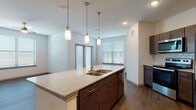 This single-floor residence offers a contemporary, open-concept layout with cohesive finishes throughout. The kitchen serves as a central focal point, showcasing a large granite-topped island with an undermount double sink and three cylindrical pendant lights suspended above. Dark espresso cabinetry lines the walls alongside stainless steel appliances, including a French door refrigerator, range, and microwave, all complemented by a white subway tile backsplash. Dark hardwood-style flooring extends through the kitchen and living area, which features a ceiling fan and access to a private exterior entry via double doors and an additional window, filling the space with natural light.

The bedrooms are finished with plush carpeting, ceiling fans, and generous windows fitted with horizontal blinds. At least one bedroom benefits from a spacious walk-in closet with built-in shelving. The bathrooms maintain the same design language found throughout the unit, with dark-framed vanity mirrors, espresso cabinetry, granite countertops, and undermount sinks. One bathroom includes wood-style tile flooring, an overhead vanity light bar, and additional wall-mounted cabinetry for storage. The overall aesthetic is clean, modern, and consistent, with neutral wall tones providing a versatile backdrop throughout the entire residence.