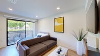 Located at 7810 Topanga Canyon Boulevard in Los Angeles, this efficiently designed one-bedroom, two-bathroom unit offers a thoughtful layout across a single floor. The open-concept living and sleeping area is bathed in natural light, courtesy of large sliding glass doors that lead to a private balcony overlooking lush greenery — a serene outdoor retreat that enhances the sense of space.

Light wood flooring runs throughout, complementing the clean white walls and recessed lighting that give the interior a bright, modern feel. The living area comfortably accommodates a full sofa and coffee table, creating a functional lounge space. The bedroom area, separated within the same open footprint, features a wall-mounted TV and a floating wood media console for a streamlined, contemporary look.

The primary bathroom is outfitted with a sleek quartz countertop vanity, an under-mount sink, warm wood cabinetry, and a backlit mirror that adds a polished, spa-like quality. The second bathroom includes a glass-enclosed shower and matching wood-toned flooring, maintaining design consistency throughout. A hallway connects the spaces efficiently, maximizing every inch of the unit. The overall ambiance is clean, modern, and welcoming — ideal for those seeking a well-appointed residence in the San Fernando Valley area of Los Angeles.