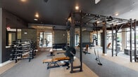 Located at 5824 McCrimmon Pkwy in Morrisville, NC, this single-floor fitness center is designed to accommodate a wide range of training styles within a generously proportioned layout. The space is divided into distinct functional zones, including a dedicated strength training area, a cardio section, and an open functional training floor.

The strength zone features heavy-duty power racks, a robust overhead rig system with gymnastic rings and battle ropes, multiple barbells with plate storage, a full dumbbell rack, and adjustable benches. The functional training area offers ample open floor space suitable for dynamic movement and conditioning work. The cardio section is equipped with treadmills, stationary bikes, and additional machines, all positioned near large windows that allow natural light and outdoor views.

Architecturally, the space features dark painted ceilings, reclaimed wood accent walls, textured stone tile panels, and a combination of carpet and hard flooring zones. Floor-to-ceiling windows and glass doors line the perimeter, maximizing natural light. Mirrored walls throughout enhance the sense of depth. The overall ambiance is modern and industrial, creating a polished yet functional training environment.