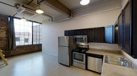 This single-floor space showcases a distinctive industrial loft aesthetic, blending raw architectural elements with modern finishes throughout. Exposed ductwork and structural beams run across the ceiling, lending an authentic urban character that is complemented by polished concrete floors and a neutral color palette. Vintage-style pendant lighting adds warmth to the industrial framework overhead.

The kitchen is a standout feature, outfitted with a full suite of stainless steel appliances including a refrigerator, electric range, built-in microwave, and dishwasher. Dark espresso cabinetry provides rich contrast against light countertops, offering ample storage and a sleek, contemporary look. The kitchen opens naturally into the main living area, reinforcing the open-concept flow of the space.

Large industrial-style windows frame views of the surrounding urban brick architecture, flooding the interior with generous natural light while connecting the space to its city context. A partial exposed brick wall further enhances the loft character, adding texture and historic charm. The overall ambiance is modern yet grounded in its industrial roots — a thoughtfully designed space that balances functionality with a bold, urban aesthetic well-suited to city living.