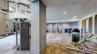 Located at 880 West Euless Boulevard in Euless, Texas, this modern two-story fitness center offers a comprehensive workout environment spread across three exercise rooms and two generously sized floors. The main level impresses with soaring double-height ceilings, floor-to-ceiling windows that flood the space with natural light, and an eye-catching motivational typography mural that anchors the room's energetic atmosphere. Geometric-patterned carpet tiles in neutral tones run throughout the strength and cardio areas, lending a contemporary aesthetic throughout.

The ground floor houses an extensive array of strength training equipment, including a power rack, cable machines, an Instinct functional trainer, a full dumbbell rack, and multiple adjustable benches. A dedicated cardio zone features rows of Star Trac treadmills positioned alongside interior windows, while a mirrored wall section expands the visual depth of the space and supports a selection of cardio machines. Circular pendant lighting accents the open areas throughout.

The upper level, accessible via an open staircase with a metal railing, provides additional workout space overlooking the main floor. A separate studio room on the upper level features light hardwood flooring, a wall-mounted television, spinning bikes, stability balls, and other group fitness accessories, making it ideal for cycling classes or guided workouts. Together, the floors create a well-rounded, professionally equipped fitness facility.