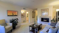 This single-floor residence offers a thoughtfully designed layout that balances comfort and functionality. The living room serves as the centerpiece of the home, featuring a tile-surround fireplace that adds warmth and a focal point to the space. Soft, neutral-toned carpeting runs throughout, complementing the warm beige walls that lend a welcoming ambiance to every room. A glass-top coffee table and carefully chosen wall art, including a vivid desert landscape painting, contribute to the space's curated, lived-in character. Natural light filters in through the windows, brightening the open interior. The living area flows seamlessly into adjoining spaces, with a hallway connecting to what appears to be a bathroom finished with light cabinetry, and access to additional rooms beyond. Ceiling vents are integrated throughout, ensuring climate comfort year-round. The overall design aesthetic is clean and understated, with warm tones and simple architectural details that create a calm, cohesive environment. Lush indoor greenery adds a natural, refreshing touch to the living area, enhancing the sense of life and vitality within the home.