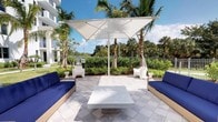 This thoughtfully designed outdoor lounge space offers a refined and comfortable setting surrounded by vibrant tropical greenery. At the heart of the space, a large cantilevered shade canopy provides generous coverage over a seating arrangement consisting of deep, cushioned sofas dressed in bold navy blue upholstery, flanking a sleek, low-profile concrete coffee table. The clean-lined furniture rests on a well-maintained stone tile patio, lending a polished, contemporary feel to the environment. Accent seating in the form of sculptural white chairs adds a modern, artistic touch to the layout. Lush landscaping frames the entire area, with tall, swaying palm trees and manicured garden beds creating a natural, resort-like backdrop. The open-air design allows for abundant natural light and fresh air circulation, enhancing the overall sense of openness and relaxation. A perimeter fence is partially visible beyond the greenery, offering a sense of privacy while maintaining a connection to the surrounding landscape. The overall ambiance is bright, airy, and inviting — ideal for casual gatherings or quiet outdoor retreats in a warm, tropical setting.