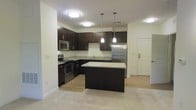 This single-floor space showcases a thoughtfully designed open-concept layout that seamlessly blends the kitchen and living areas. The kitchen is a standout feature, boasting rich dark espresso cabinetry that contrasts elegantly with light granite countertops. Stainless steel appliances, including a refrigerator, range, and microwave, lend a polished, contemporary feel. A central kitchen island with a matching granite surface provides additional workspace and a natural gathering point. Above the island, two sleek pendant lights hang at varying heights, adding a stylish focal point, while flush-mount ceiling fixtures ensure the entire space is well-illuminated. The flooring transitions from warm light hardwood in the kitchen to plush carpet in the adjacent living area, defining each zone while maintaining a cohesive aesthetic. Clean white walls and a neutral color palette throughout amplify the sense of openness and brightness. An entry door with a modern handle is visible to the side, reinforcing the functional flow of the layout. Overall, the space presents a clean, modern ambiance that balances practicality with refined design details.