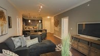 This single-floor residence offers a thoughtfully designed open-concept layout that effortlessly connects the main living areas, creating a cohesive and inviting atmosphere. The living room serves as the centerpiece of the space, featuring rich dark hardwood flooring, crown molding, and warm neutral-toned walls that lend a modern yet comfortable character. A spacious sectional sofa anchors the living area, which flows naturally into the dining space, complete with a round dining table and cushioned chairs ideal for entertaining.

The kitchen is well-appointed with dark cabinetry, stainless steel appliances, and a kitchen island that doubles as a breakfast bar. Pendant lighting above the island and track lighting along the ceiling add layers of illumination, enhancing both functionality and ambiance throughout the open space.

Crisp white trim and panel doors provide a clean contrast against the darker tones used throughout. Decorative wall art and thoughtful accessory placement contribute to the curated feel of the interior. A rustic wooden dresser near the living area adds a touch of warmth and texture.

Overall, this residence balances contemporary design with warm, livable elements, making it a welcoming and visually cohesive space suited for both everyday comfort and stylish entertaining.