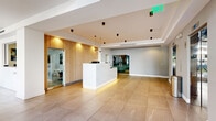 Located on Wilshire Boulevard in Los Angeles, this elegantly designed lobby and lounge space offers a refined, contemporary aesthetic throughout its single-floor layout. The reception area serves as the centerpiece, featuring a striking marble-clad desk with integrated underlighting, set against a warm vertical wood slat accent wall that adds texture and sophistication. Large-format stone tiles extend across the floor, unifying the space with a clean, polished finish.

The hallway connects the reception to adjacent areas seamlessly, maintaining a consistent design language of crisp white walls, recessed lighting, and modern black cylindrical light fixtures mounted to the ceiling. Glass-paneled doors and partitions introduce transparency and openness throughout.

The living lounge area is bathed in abundant natural light, thanks to expansive floor-to-ceiling windows with slim black frames that frame lush outdoor greenery and views of the surrounding Westside neighborhood. Tasteful furnishings — including neutral-toned lounge chairs, a low glass coffee table, and curated indoor plants — contribute to the space's calm, welcoming ambiance. A statement black articulating wall sconce adds a designer touch overhead.

Together, the spaces create a polished, minimalist environment that balances functionality with refined architectural detail, ideal for welcoming guests in a professional setting.