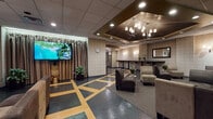 Located at 12 Park Place in Hartford, CT, this single-floor lounge space offers a sophisticated and well-appointed interior designed for comfort and social interaction. The space is anchored by a striking coffered ceiling section finished in deep bronze tones, from which a multi-arm chandelier with lamp-style shades hangs as a central focal point, complemented by recessed lighting throughout.

The flooring is a standout architectural feature, combining dark slate-toned tiles with bold geometric gold inlay patterns that guide movement through the space and add visual depth. Textured, paneled wall treatments in warm neutral tones create a refined backdrop across the entire floor.

Seating is arranged in flexible clusters featuring plush, upholstered chairs and sofas in earthy taupe and charcoal tones, accented by metallic side tables. Framed artwork is displayed at multiple points along the walls, adding a gallery-like quality to the ambiance. Wall-mounted sconces provide warm, ambient lighting throughout.

A bar-height counter with stools runs along one side, suitable for casual mingling or dining. Draped floor-to-ceiling curtains along one wall add softness and a sense of privacy. A large flat-screen television is mounted near the curtained area, enhancing the space's versatility for events or presentations. The overall atmosphere is polished, welcoming, and well-suited for gatherings or receptions.