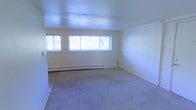 This single-floor space features an open living area finished with wall-to-wall carpeting in a neutral tone, contributing to a clean and understated aesthetic. The room is generously proportioned, offering a sense of openness that lends itself well to flexible furniture arrangements. A row of horizontal windows along one wall allows natural light to filter in, brightening the cream-colored walls and creating an airy atmosphere. Beneath the windows, a baseboard heating system runs the length of the wall, providing an efficient and unobtrusive heating solution. The flat, smooth ceiling complements the minimalist design of the space. The overall palette is consistent and neutral throughout, with white walls and light carpeting creating a cohesive, calm environment. The layout appears straightforward and functional, with clear sightlines across the room. The space is currently vacant, making it easy to envision a variety of interior configurations. Overall, the room presents a simple, well-lit interior that offers a practical and adaptable living environment.