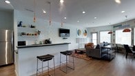 This single-floor space showcases a thoughtfully designed open-plan layout that seamlessly blends a kitchenette and lounge area into one cohesive environment. The kitchenette features a sleek dark countertop bar with modern bar stools, a small sink, and a coffee station, backed by a striking textured white tile accent wall that adds depth and visual interest. Three pendant lights with exposed Edison bulbs hang from orange cords, contributing an industrial-chic aesthetic that complements the recessed lighting throughout.

The lounge area is comfortably furnished with a leather armchair and a large sofa arranged around a colorful area rug, creating an inviting gathering space. A wall-mounted flat-screen TV is centrally positioned for easy viewing from both the bar and seating areas. A white modular shelving unit provides practical storage and display space, while two framed circular art pieces add a decorative accent to the walls. Warm wood-look flooring runs throughout, tying the space together, and a window along the far wall allows natural light to filter in, enhancing the bright, airy atmosphere.