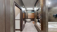 Located at 246–276 Kyser Blvd in Madison, Alabama, this well-appointed commercial suite offers a refined and functional layout across a single floor. The space is designed with a distinctive aesthetic, featuring deep charcoal faux-finish walls, dark-trimmed doors, and large framed mirrors that lend a sophisticated, upscale atmosphere throughout.

The central corridor serves as the spine of the space, lined with private room entrances on both sides and anchored by a round upholstered ottoman and decorative area rug at its end. Warm wood-toned roller blinds filter natural light through transom windows, complementing the rich, moody interior palette. Light tile flooring runs consistently throughout, providing a clean contrast to the darker wall tones.

Three bathrooms are included in the suite, each finished with granite-topped dual vanities, large wall mirrors, modern dispensers, and private stall partitions — reflecting a commercial-grade standard of function and cleanliness. A dedicated tanning room houses a full-length tanning bed and includes wall-mounted supply storage and a small seating bench, offering a private and efficient client experience.

The entryway features warm neutral tones, decorative floral artwork, and an arched-panel door that sets an inviting tone upon arrival. This space is ideally suited for a beauty, wellness, or personal care business seeking a polished, client-ready environment.