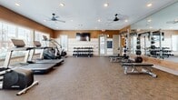 Located at 120 SR-76 in White House, Tennessee, this fitness center offers a thoughtfully designed, single-floor layout that provides ample space for a full range of workout activities. The main gym area features carpet flooring and is outfitted with a wide array of equipment, including treadmills, rowing machines, cable machines with TRX suspension systems, and a Hammer Strength power rack with an adjustable bench. A dedicated free weights section includes a full dumbbell rack and kettlebell sets, complemented by a wall-mounted flat-screen TV for entertainment. Dual ceiling fans and recessed lighting keep the space well-ventilated and brightly lit, while large windows allow natural light to fill the room and offer views of the surrounding area. Warm earth-tone accent walls and wood-paneled feature walls lend a modern yet welcoming aesthetic throughout. A connected studio room, accessible through a glass-panel door, provides a separate area for flexibility, stretching, or small-group training. This secondary space features floor-to-ceiling mirrors, a ceiling fan, its own recessed lighting, and a mounted TV, creating a versatile environment for a variety of fitness disciplines. Together, both areas form a comprehensive, well-appointed fitness facility designed to support diverse exercise routines.