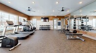 Located at 120 SR-76 in White House, Tennessee, this fitness center offers a thoughtfully designed, single-floor layout that provides ample space for a full range of workout activities. The main gym area features carpet flooring and is outfitted with a wide array of equipment, including treadmills, rowing machines, cable machines with TRX suspension systems, and a Hammer Strength power rack with an adjustable bench. A dedicated free weights section includes a full dumbbell rack and kettlebell sets, complemented by a wall-mounted flat-screen TV for entertainment. Dual ceiling fans and recessed lighting keep the space well-ventilated and brightly lit, while large windows allow natural light to fill the room and offer views of the surrounding area. Warm earth-tone accent walls and wood-paneled feature walls lend a modern yet welcoming aesthetic throughout. A connected studio room, accessible through a glass-panel door, provides a separate area for flexibility, stretching, or small-group training. This secondary space features floor-to-ceiling mirrors, a ceiling fan, its own recessed lighting, and a mounted TV, creating a versatile environment for a variety of fitness disciplines. Together, both areas form a comprehensive, well-appointed fitness facility designed to support diverse exercise routines.
