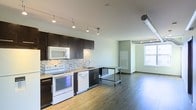 This single-floor space showcases a thoughtfully designed open-concept layout that seamlessly connects the kitchen and living areas. The kitchen is a standout feature, outfitted with dark espresso cabinetry that contrasts beautifully against a mosaic tile backsplash in neutral tones. White appliances, including a range, microwave, and dishwasher, are neatly integrated beneath the cabinetry, complemented by a sleek countertop and an undermount sink. Track lighting runs along the ceiling, providing focused illumination throughout the kitchen area.

Industrial character is introduced through an exposed ventilation duct that runs along the upper portion of the space, adding a loft-like aesthetic. Durable hardwood-style flooring extends throughout, lending warmth and continuity across the open floor plan. A large window in the adjacent living area floods the space with natural light, creating a bright and welcoming atmosphere. The open layout between the kitchen and living zone offers a flexible, functional arrangement well-suited for everyday living and entertaining.