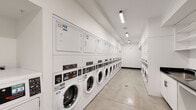 This shared laundry facility is designed for high-capacity use, offering a clean, functional, and efficiently organized environment. The main laundry area features an extensive row of front-loading washing machines paired with stacked dryers lining the walls, providing ample capacity for multiple users simultaneously. The polished concrete flooring throughout the space is both durable and easy to maintain, contributing to the facility's practical aesthetic.

The space is finished with crisp white walls and illuminated by linear LED lighting that runs along the ceiling, ensuring bright, consistent visibility throughout. A built-in cabinetry station with a dark countertop and utility sink provides a convenient area for hand-washing or pre-treating garments, while open white shelving above offers organized storage for laundry supplies. An additional stainless steel worktable is positioned in a secondary alcove area, serving as an extra folding or sorting surface.

Climate comfort is addressed with a wall-mounted split air conditioning unit, keeping the room pleasant during use. Service information and laundry app instructions are displayed on the wall, indicating a modern, app-enabled payment and monitoring system. The overall design prioritizes functionality, cleanliness, and user convenience, making this a well-appointed communal laundry space suited for a residential building or similar multi-user facility.