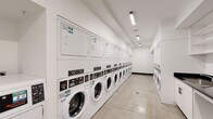 This shared laundry facility is designed for high-capacity use, offering a clean, functional, and efficiently organized environment. The main laundry area features an extensive row of front-loading washing machines paired with stacked dryers lining the walls, providing ample capacity for multiple users simultaneously. The polished concrete flooring throughout the space is both durable and easy to maintain, contributing to the facility's practical aesthetic.

The space is finished with crisp white walls and illuminated by linear LED lighting that runs along the ceiling, ensuring bright, consistent visibility throughout. A built-in cabinetry station with a dark countertop and utility sink provides a convenient area for hand-washing or pre-treating garments, while open white shelving above offers organized storage for laundry supplies. An additional stainless steel worktable is positioned in a secondary alcove area, serving as an extra folding or sorting surface.

Climate comfort is addressed with a wall-mounted split air conditioning unit, keeping the room pleasant during use. Service information and laundry app instructions are displayed on the wall, indicating a modern, app-enabled payment and monitoring system. The overall design prioritizes functionality, cleanliness, and user convenience, making this a well-appointed communal laundry space suited for a residential building or similar multi-user facility.