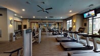 Located at 1916 W Gray St in Houston's River Oaks area, this well-appointed fitness center offers a comprehensive workout environment across a single, open floor plan. The space is designed to accommodate a full range of exercise needs, combining dedicated cardio and strength training zones in a cohesive layout.

The cardio area is lined with multiple Life Fitness treadmills and elliptical machines positioned along large floor-to-ceiling windows that provide natural light and views of the surrounding outdoor pool area. Mounted flat-screen TVs are strategically placed throughout, keeping users entertained during their workouts.

The strength training section features a multi-station cable machine, adjustable benches, a dumbbell rack, and an assortment of medicine balls displayed on a dedicated rack. Motivational workout instruction posters line the walls, adding a functional and visual element to the space.

Large wall-to-wall mirrors in the strength zone enhance the sense of openness and allow users to monitor their form. The flooring throughout consists of durable, patterned carpet tiles suited for gym use. Ambient lighting is provided by a combination of recessed ceiling lights and warm wall sconces, while ceiling fans help maintain air circulation across the space. The overall atmosphere is modern, functional, and well-maintained.