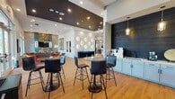 Located at 2571 Seminole Trail in Charlottesville, VA, this single-floor community clubhouse is a thoughtfully designed gathering space that blends contemporary style with welcoming functionality. The open-plan layout flows seamlessly between distinct zones, each finished with warm hardwood flooring and carefully curated décor.

The main lounge area is anchored by a striking accent wall of stacked stone and bold color-blocked paneling, complemented by a sophisticated mix of leather and upholstered seating arranged around a modern coffee table. Soaring ceilings, oversized windows, and a dramatic geometric chandelier create an airy, light-filled atmosphere with panoramic outdoor views of the surrounding landscape.

A casual café-style seating area features high-top bistro tables with sleek bar stools, positioned alongside a built-in wet bar with light-toned cabinetry and a decorative tile backsplash accented by geometric wall sconces.

The kitchen is fully appointed with gray shaker-style cabinetry, dark quartz countertops, stainless steel appliances, a matte black faucet, and a decorative patterned tile floor. A large central island with upholstered barstools provides ample counter seating beneath a trio of angular brass pendant lights.

A dedicated dining area offers an expansive oval table surrounded by modern chairs, bathed in natural light from floor-to-ceiling windows framed by exposed brick, completing this refined and versatile community space.