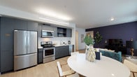 This single-floor residence offers a thoughtfully designed open-concept layout that seamlessly connects the kitchen, dining, and living areas into one cohesive living space. The kitchen is outfitted with stainless steel appliances — including a full-size refrigerator, electric range with oven, built-in microwave, and dishwasher — complemented by sleek dark grey cabinetry and a clean white backsplash. A linear pendant light fixture overhead adds a modern industrial touch while ensuring ample task lighting. The dining area features a round white table with contemporary chairs, creating an inviting space for meals and gatherings. Beyond the dining area, the living space is highlighted by a deep navy accent wall that adds depth and contrast to the otherwise light, neutral palette. Light wood-tone flooring runs throughout, contributing to a warm and airy atmosphere. Lush indoor plants add natural texture and a refreshing pop of greenery. The layout flows openly toward additional rooms, suggesting generous connectivity throughout the residence. The overall design aesthetic is clean, modern, and functional, making excellent use of the available space.