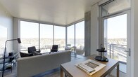 This single-floor space is defined by its dramatic floor-to-ceiling corner windows, which flood the interior with natural light and offer expansive panoramic views of the cityscape and waterfront beyond. The open-plan layout seamlessly connects the dining and living areas, creating a cohesive and airy environment. Exposed concrete columns add an industrial architectural contrast to the bright white ceiling and light-toned flooring, lending the space a contemporary, urban character. The living area is furnished with a neutral-toned sofa and a curated arrangement of accent chairs and side tables, oriented to take full advantage of the sweeping views. A dining table positioned near the windows is set with place settings, reinforcing the space's functional elegance. The corner window configuration maximizes sightlines in multiple directions, making natural light a constant presence throughout the day. The overall ambiance is sleek and modern, balancing minimalist design sensibilities with warm, livable furnishings, resulting in a sophisticated environment suited to both relaxing and entertaining at considerable elevation above the surrounding city.