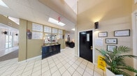 This single-floor community space is designed to provide a warm and functional environment for residents and visitors alike. The entry foyer sets an inviting tone with light-toned tile flooring, a decorative black sideboard with a gilded mirror, and a distinctive glass block window partition that allows diffused natural light to filter throughout. Elevated ceilings in the entry area transition into dramatically vaulted ceilings supported by prominent white structural columns in the main lounge, enhancing the sense of openness and grandeur. The central gathering area features plush seating arranged around a coffee table atop carpeted flooring, ceiling fans for comfort, and olive-toned accent walls that lend warmth to the neutral palette. A dedicated seating nook along the perimeter is bathed in natural light from large picture windows that frame views of the landscaped exterior and outdoor pool area. A dining area with a dark wood table and chairs sits beneath expansive windows, providing bright, garden-facing seating. A workstation area with dark furnishings is visible beyond the entry, suggesting a leasing or administrative function. Wall sconces and table lamps throughout contribute to a layered, welcoming lighting scheme that complements the space's community-focused design.