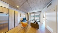 This contemporary 950 square foot apartment, located in the heart of downtown, showcases a perfect blend of industrial and modern design elements. The space features two bedrooms, including a spacious 320 square foot primary bedroom, and one bathroom. The apartment's most striking feature is its exposed concrete ceilings, which add an urban, loft-like atmosphere to the entire space.

Large windows dominate the living areas, flooding the apartment with natural light and offering views of the surrounding cityscape. The open-concept living and kitchen area is ideal for entertaining, with a sleek, modern kitchen featuring two-tone cabinetry, stainless steel appliances, and ample counter space.

The bedrooms continue the minimalist aesthetic with clean lines and built-in storage solutions. Hardwood floors run throughout the apartment, adding warmth to the industrial elements. The bathroom, while not pictured, likely follows the same modern design principles.

A small balcony accessible from the living room provides outdoor space, perfect for enjoying morning coffee or evening relaxation. The apartment's location in a multi-story building with large windows and brick exterior suggests a vibrant, urban setting.

Overall, this apartment offers a stylish, contemporary living space that maximizes its 950 square feet through clever design and an emphasis on openness and light.