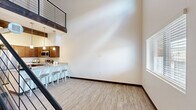 Located at 1571 North Pine Cliff Drive in Flagstaff, Arizona, this two-story loft-style space offers a thoughtfully designed layout spread across two floors. The main level serves as the heart of the space, highlighted by dramatic soaring ceilings that create an impressive sense of volume and openness. Light wood-look flooring runs throughout, complementing the clean, neutral palette of the walls. The open-concept kitchen features rich walnut-toned cabinetry, stainless steel appliances, and a granite-topped peninsula with bar seating—ideal for casual dining and entertaining. Pendant lighting adds a warm, modern touch above the kitchen area. A bold, open-riser staircase with dark wood treads and sleek black metal cable railings serves as a striking architectural centerpiece, leading to the upper loft level. The loft overlooks the main floor through matching metal railings, offering an airy, connected feel between the two levels. Natural light filters in through well-placed windows on both floors, brightening the interior throughout the day. The bathroom is well-appointed with rich wood cabinetry, a granite countertop, a large vanity mirror, and clean, neutral finishes. This space combines modern industrial design elements with warm, inviting materials for a distinctive and functional living environment.