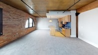 This single-floor loft unit showcases a compelling mix of industrial heritage and contemporary updates. The open-concept living and kitchen area is the heart of the space, distinguished by striking exposed brick walls, warm wood-plank ceilings, and rail track lighting that runs overhead. A large arched window floods the main living area with natural light, highlighting the soft carpeted flooring and the expansive, airy feel of the room.

The kitchen is thoughtfully designed with light maple cabinetry, a bold navy blue accent wall, stainless steel appliances including a refrigerator, electric range with over-the-range microwave, and a built-in dishwasher. A freestanding kitchen island with a matching maple finish provides additional workspace and serves as a natural divider between the kitchen and living areas. Original heavy timber columns punctuate the space, reinforcing the building's historic character.

Two additional carpeted rooms offer flexible use as bedrooms or office spaces, each featuring the same wood-plank ceiling aesthetic. Two bathrooms serve the unit: a full bath with a tub-shower combination, warm wood vanity, and tile flooring, and a second bath with a glass-enclosed walk-in shower, similarly finished with tile flooring and a wood vanity. Together, these elements create a refined yet character-rich living environment.