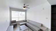 This single-floor living space offers a clean, minimalist design with a neutral color palette throughout. The main living area is furnished with a contemporary grey sectional sofa and matching accent chair, complemented by a low-profile rectangular coffee table with a sleek metal frame. Light wood-tone flooring runs throughout, enhancing the bright, airy atmosphere of the room. A large window fitted with horizontal blinds allows generous natural light to filter in, offering views of the surrounding outdoor greenery and neighboring buildings. A five-blade ceiling fan with an integrated light fixture is centrally mounted, providing both ventilation and ambient lighting. The white walls and minimal decor contribute to an uncluttered, functional aesthetic. The overall layout is efficiently organized, making practical use of the available space while maintaining an open and comfortable feel suited to everyday living.