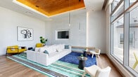 Located at 1747 Van Buren St in Hollywood, Florida, this single-floor community lounge is a thoughtfully designed gathering space that blends modern aesthetics with a welcoming, resort-inspired atmosphere. The open-plan layout encompasses a generous living area, a social dining zone, and a stylish bar counter, all unified by warm light wood flooring that runs throughout.

The living area serves as a comfortable focal point, anchored by a large sectional sofa in neutral tones, vibrant yellow accent chairs, and colorful striped area rugs that inject energy into the space. A wall-recessed flat-screen television and a warm wood-paneled tray ceiling with ambient lighting lend a refined, residential feel.

The dining and bar zone features a long communal table with upholstered seating, pendant lighting, and a wet bar accented by an industrial-style steel shelving structure adorned with lush greenery. White quartz countertops and bar stools complete this social hub.

Floor-to-ceiling glass walls and double glass entry doors flood the interior with abundant natural light while offering sweeping views of the manicured outdoor lawn, palm trees, and communal seating areas beyond. The seamless indoor-outdoor connection is a defining characteristic of this space, making it ideal for relaxed socializing or community events in the heart of Hollywood, FL.