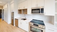 This efficiently designed single-floor studio offers a clean, contemporary aesthetic throughout, characterized by crisp white walls, light hardwood-style flooring, and recessed lighting that enhances the bright, airy ambiance. The open living area is anchored by large floor-to-ceiling windows that flood the space with natural light and provide views of the surrounding urban landscape, with a sliding glass door leading to a private balcony. A wall-mounted HVAC unit ensures year-round comfort. The kitchen is a standout feature, outfitted with a full suite of stainless steel appliances including a refrigerator, electric range, over-the-range microwave, and dishwasher. White flat-front cabinetry extends to the ceiling and is complemented by warm wood open shelving, light quartz countertops, and a classic white subway tile backsplash, creating a polished and functional cooking space. The bathroom maintains the same modern, minimal palette with a white vanity offering under-sink storage, a toilet, and a glass-enclosed shower. Towel bars in brushed nickel add a refined finishing touch. Overall, the space presents a cohesive, well-appointed interior that maximizes functionality without sacrificing style.