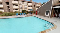 This amenity space features two distinct areas designed to support an active lifestyle: an outdoor swimming pool and an indoor fitness center. The pool area is generously sized with a curvilinear shape and clear blue water. A spacious pool deck surrounds the water, furnished with blue lounge chairs for relaxation. A shaded seating area with a blue umbrella and table provides a comfortable retreat. Lush landscaping lines the perimeter, and the multi-story residential building backdrop creates an enclosed, community-oriented atmosphere.

The fitness center is a well-lit, single-floor space distinguished by bold orange and blue accent stripes along the upper walls, lending the room an energetic character. Large windows along one wall overlook the pool area, flooding the interior with natural light. The space is carpeted with a patterned tile-style flooring and equipped with an array of cardio machines, including multiple treadmills and an elliptical trainer. Strength-training options include a multi-function cable machine, stationary bikes, a rowing machine, and a full dumbbell rack backed by a large wall mirror. A ceiling fan and recessed lighting ensure comfort throughout workouts.